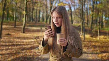 Side view of stylish, beautiful lady walking in park in autumn. Attractive, young female standing, using smartphone, happy, surprised, looking at camera. Concept of happiness.