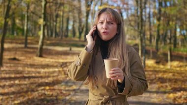 Side view of attractive, young woman standing in park, talking on phone. Fashionable lady wearing trench, quarreling, angry, drinking coffee. Concept of urban lifestyle.