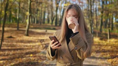 Side view of fashionable, young lady walking, standing in park, enjoying, smiling. Attractive, cute woman using smartphone, drinking coffee. Concept of modern lifestyle.
