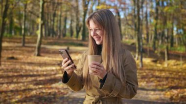 Side view of positive, smiling lady using smartphone, scrolling, reading, texting. Attractive, young woman standing in park, drinking coffee. Concept of modern lifestyle.