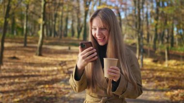 Front view of positive, happy lady standing in park, using smartphone, scrolling. Attractive woman wearing stylish trech, holding paper cup, drinking coffee. Concept of modern lifestyle.