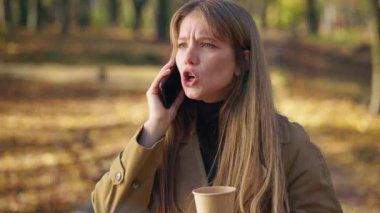 Side view of angry, young lady talking on smartphone in park. Attractive girl standing, using smartphone, drinking coffee, holding paper cup. Concept of modern lifestyle.