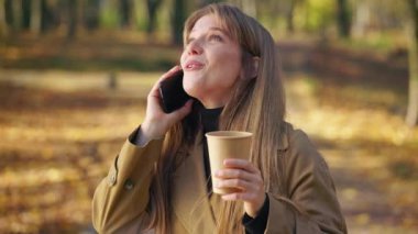 Side view of pretty, young female talking on phone in park in autumn. Attractive woman wearing stylish clothes, smiling, standing, drinking coffee. Concept of modern lifestyle.