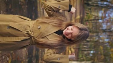 Front view of positive, smiling lady walking in park in autumn, standing. Cheerful woman looking at camera, raising hands, showing hurray. Concept of happiness and enjoyment.