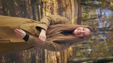 Front view of elegant, cheerful woman standing, walking in park in autumn. Young lady looking at camera, smiling, raising hands. Concept of happiness and sincere emotions.