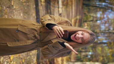 Front view of smiling woman standing in park, using smartphone. Elegant lady scrolling, reading, surprised, shocked., opening mouth. Concept of modern technologies and lifestyle.