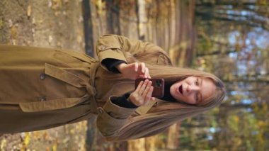 Front view of elegant, stylish woman walking, standing in park in autumn. Glad, cheerful lady using smartphone, reading, surprised, opening mouth. Concept of sincere emotions.