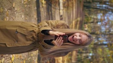 Front view of elegant lady wearing stylish clothes, walking, standing in park. Cheerful woman using smartphone, reading, surprised, opening mouth. Concept of happiness and joy.