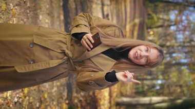 Front view of elegant, young lady standing in park, using smartphone. Happy, glad female wearing stylish coat, scrolling, smiling. Concept of modern technologies and lifestyle.