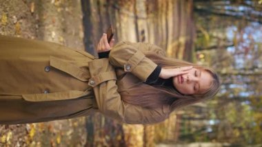 Front view of young woman standing in park, using smartphone, having headache. Pretty, glad lady wearing stylish clothes, scrolling, nervous. Concept of modern urban lifestyle.