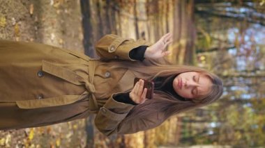 Front view of tired, disappointed, nervous woman standing in park, using smartphone. Beautiful lady wearing stylish clothes, Concept of modern technologies and lifestyle.