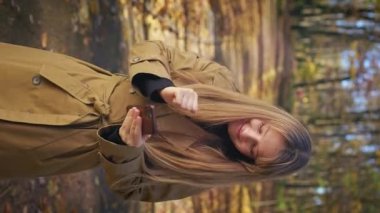 Front view of positive, cheerful woman walking in park, enjoying autumn weather. Pretty, cute lady using smartphone, dancing, closing eyes. Concept of happiness and enjoyment.