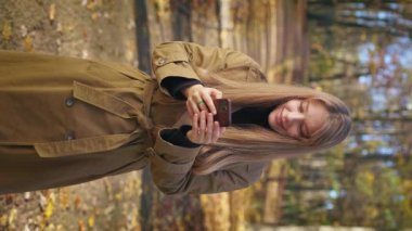 Front view of surprised, shocked lady standing in park, using smartphone. Cheerful, young woman scrolling, texting, opening mouth. Concept of modern technologies and living.
