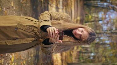 Front view of happy, glad female walking in park in autumn. Cheerful lady using smartphone, reading scrolling, smiling, admiring, enjoying. Concept of happiness and harmony.