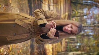 Front view of angry, furious lady standing in park, holding using smartphone. Pretty, elegant woman wearing stylish clothes, pressing button. Concept of modern urban lifestyle.