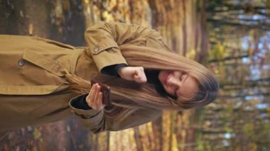 Front view of attractive, cheerful lady walking, standing in park in autumn. Happy, glad female wearing stylish clothes, using smartphone, dancing. Concept of modern lifestyle.