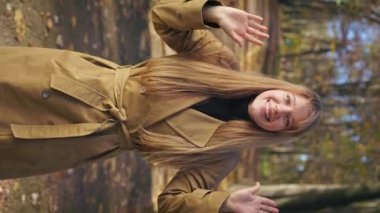 Front view of elegant, stylish woman walking in park in autumn, standing. Pretty, glad lady raising hands, waving, smiling, looking at camera. Concept of happiness and sincere emotions.