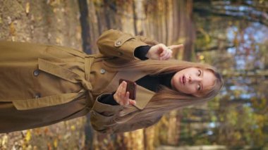 Side view of attractive, young woman walking in park in autumn, standing. Happy, glad lady holding, using smartphone, thinking, remembering. Concept of modern urban lifestyle.