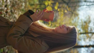 Side view of attractive, elegant woman walking, standing in park. Happy, glad lady holding paper cup, drinking hot coffee, smelling, enjoying. Concept of happiness and enjoyment.