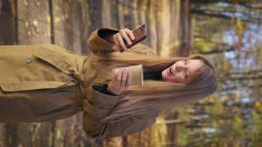 Side view of positive, cheerful lady using smartphone, reading, surprised, pleased. Happy, glad woman walking in park, holding coffee. Concept of modern technologies and lifestyle.