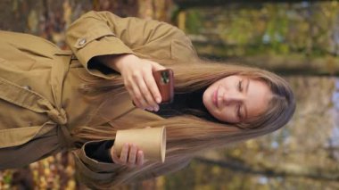 Front view of surprised, young lady using smartphone in park, looking at camera. Cheerful, elegant woman holding paper cup, drinking coffee. Concept of happiness and sincere emotions.