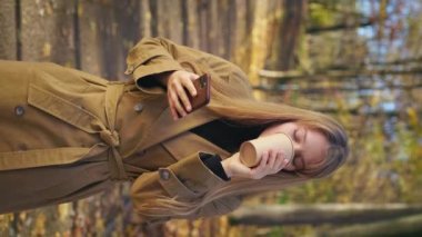 Side view of cheerful, happy woman walking in park in autumn, standing. Beautiful, young lady standing, using smartphone, drinking coffee. Concept of happiness and enjoyment.
