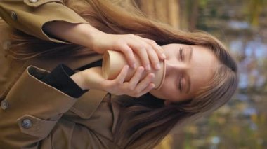 Side view of pretty, cute lady walking, enjoying sunny autumn weather. Attractive, young woman holding paper cup, drinking coffee. Concept of happiness and harmony with nature.