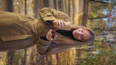 Side view of elegant lady walking in park in sunny autumn weather, standing. Attractive, young woman using smartphone, drinking coffee, admiring. Concept of harmony with nature.