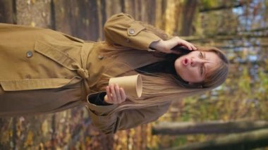Side view of angry lady talking on smartphone in park in autumn. Beautiful woman quarreling, holding paper cup, drinking coffee outdoors in fall. Concept of modern lifestyle.