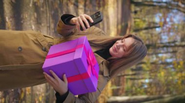Side view of glad, happy, satisfied standing in park, holding present, gift box. Beautiful lady using smartphone, scrolling, smiling. Concept of sincere emotions and happiness.