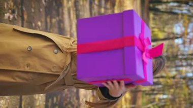 Front view of cheerful, cute lady holding present, gift box in park. Beautiful, young female wearing stylish coat, holding present, gift box, giving. Concept of sincere emotions.