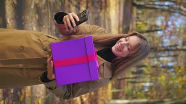 Side view of happy, elegant lady wearing stylish clothes, walking in park. Beautiful, young female holding present, gift box, using smartphone. Concept of happiness and enjoyment.