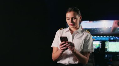Front view of pretty, female pilot stewardess having break, standing in cabin. Woman wearing uniform, using smartphone, reading, smiling. Concept of aviation and airplane.