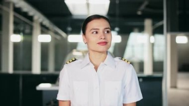 Front view of young, beautiful woman working as pilot instructor. Female pilot wearing uniform, walking in hall, looking asides, smiling, happy, glad. Concept of aviation tourism.