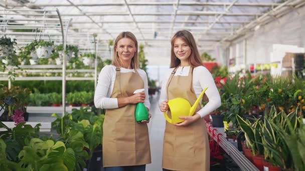 两个年轻快乐的女士穿着制服在温室里工作漂亮的女人看着相机笑着站着种植和照料植物的概念 — 图库视频影像