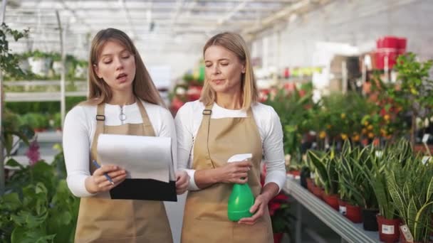 两个女花匠站在温室的侧视图 华丽的 漂亮的女人穿着制服拿着文件夹照顾植物的概念 — 图库视频影像