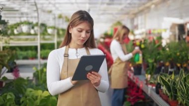 Üniforma giyen, ayakta duran, serada çalışan çekici bir kadın. Genç çiçekçi elinde tablet, not alıyor, daktilo ediyor, kaydırıyor. Modern yaşam tarzı kavramı.