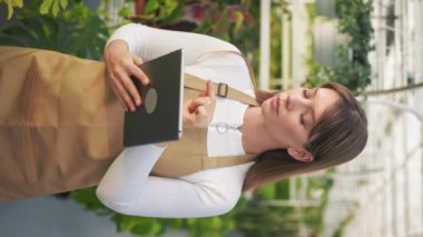 Genç bayan çiçekçinin serada dikilip bitkileri saymasının yan görüntüsü. Çekici bir kadın tutuyor, tablet kullanıyor, not alıyor. Büyüme ve bitkilerle ilgilenme kavramı.