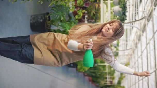 侧视图年轻的 女性花匠浇灌 在温室喷洒植物 漂亮的女孩穿着围裙 拿着瓶子 照顾植物的概念 — 图库视频影像