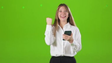 Stüdyoda çalışan, poz veren, mutlu, neşeli bir bayanın ön görüntüsü. Güzel, genç bir kadın akıllı telefon kullanıyor, yaşasın diyor, gülümsüyor. Yeşil stüdyo arka planında izole edilmiş..