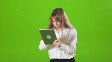 Genç, sarışın, resmi kıyafetlerin yan görüntüsü, stüdyoda ayakta, poz verirken. Öfkeli, sinirli bir kadın elinde tablet tutuyor, bastırıyor. Yeşil stüdyo arka planında izole edilmiş..