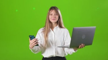 Resmi kıyafetler giyen, elinde akıllı telefon ve dizüstü bilgisayarla genç sarışın bir kadının ön görüntüsü. Güzel, neşeli bayan kameraya bakıyor. Yeşil stüdyo arka planında izole edilmiş..