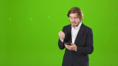 Kızgın, sinirli bir adamın stüdyoda dikilip, elinde tuttuğu, akıllı telefon kullandığı, daktilo kullandığı görüntüler. Kostüm ve gömlek giyen yakışıklı bir iş adamı. Yeşil stüdyo arka planında izole edilmiş..
