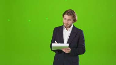Resmi kıyafetler giyen, klasör tutan, yazı yazan işadamlarının ön görüntüsü. Yakışıklı, genç bir erkek ofiste çalışıyor, ayakta duruyor, düşünüyor. Yeşil stüdyo arka planında izole edilmiş..