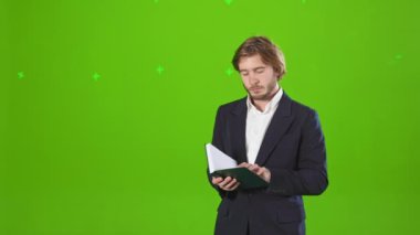Kostüm giymiş, ayakta duran, stüdyoda poz veren yakışıklı bir erkek. Sakallı bir adam elinde defter, okuyor, ciddi, meşgul. Yeşil stüdyo arka planında izole edilmiş..