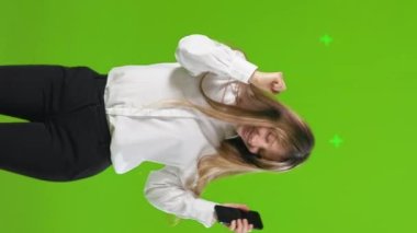 Akıllı telefonlu, dans eden, stüdyoda eğlenen mutlu genç bir kadın. Heyecanlı, tişörtlü, cep telefonuyla müzik dinleyen, yeşil arka planda telefonlarını kontrol eden güzel bir kız öğrenci. Duygular konsepti.