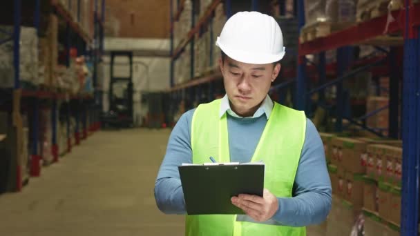 专业的亚洲主管 身穿白色硬帽和反光背心 在大型工厂仓库对产品进行盘点 男子在剪贴板上重点记录货架上的用品 — 图库视频影像