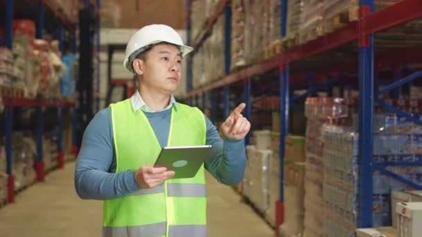 亚洲仓库工人站在宽敞的仓库中 数着货架上的供应品 以现代数码平板电脑管理制服的成熟男子为重点 — 图库视频影像