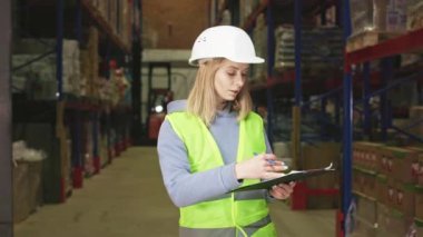 Depo envanteri sırasında panodaki belgelere bakan, sarı saçlı, konsantre bir kadın yönetici. İş yerinde beyaz şapka ve yansıtıcı yelek giyen beyaz bir genç kadın..