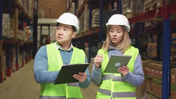 白人妇女和亚洲男子与平板和剪贴板 计数和检查货物在仓库货架 戴安全帽的有重点的同事把信息放到公司应用程序上 — 图库视频影像
